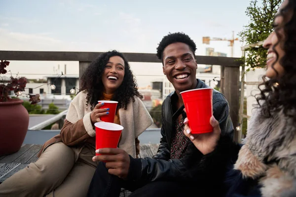 Grupo de jóvenes adultos multiculturales riéndose entre sí, sentados en una terraza en la azotea socializando, bebiendo — Foto de Stock