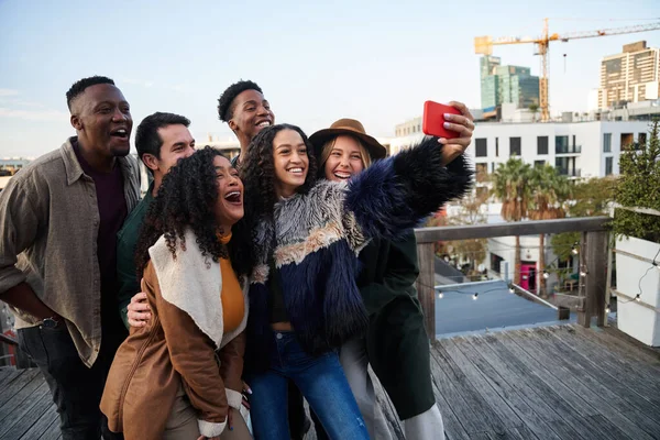 Groupe multiculturel d'amis prenant un selfie lors d'une fête sur le toit. Riant à des visages drôles, fond de paysage urbain. — Photo
