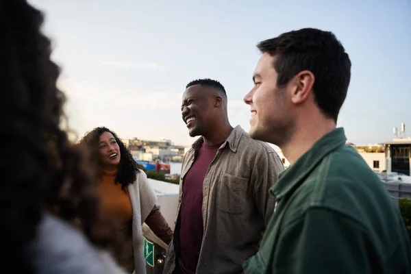 Группа мультикультурных друзей смеются и общаются на террасе на крыше в сумерках. — стоковое фото