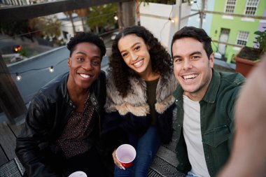 Selfie çekerken gülümseyen bir grup çok kültürlü genç yetişkin. Şehirdeki bir çatı partisinde, güzel zamanlardı..