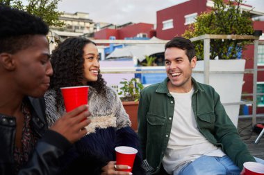 Bir grup çok kültürlü yetişkin arkadaş birlikte şakaya gülüyorlar. Şehirdeki bir çatı partisinde oturmak, güzel zamanlardı..