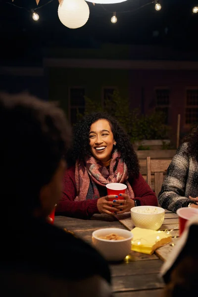 Biracial jeune adulte femelle socialisant à une table sur le toit. Assis à table avec des amis, tenant une tasse rouge, riant — Photo