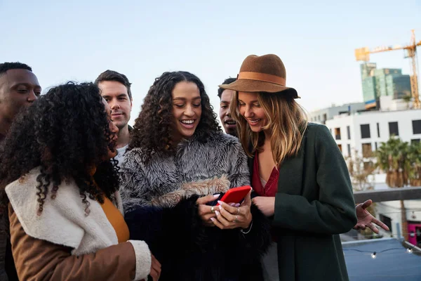 Diverso grupo de amigos adultos jóvenes en una terraza en la azotea de la ciudad, mirando y riéndose de una foto de niñas biraciales teléfono celular. —  Fotos de Stock