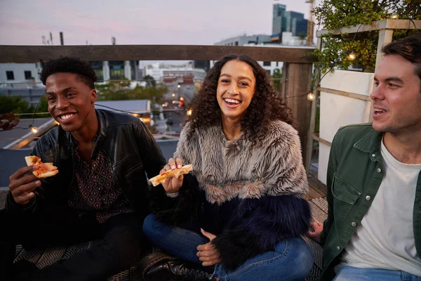 Group of multi-cultural young adult friends sitting at rooftop party eating pizza in the city.