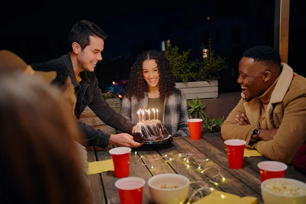 Kaukasischer Mann legt Kuchen für birassisches Geburtstagskind hin. Gruppe unterschiedlicher junger Erwachsener sitzt bei Dachparty am Tisch und feiert Geburtstagskind mit Kuchen. — Stockfoto