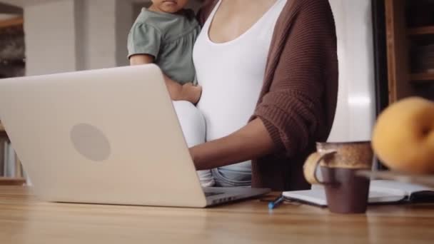 Trabajo desde casa madre biracial sosteniendo a su hija pequeña en su brazo mientras trabaja en una cocina de estilo moderno — Vídeos de Stock