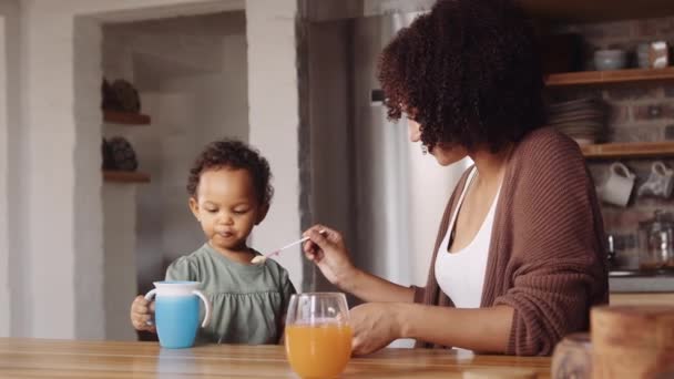 Biracial mother feeding her young daughter food in modern-styled kitchen. Child grabbing cup. — Stock Video
