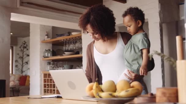 Biracial matka pracuje na laptopie z domu ze swoją młodą córką w ramię w nowoczesnej kuchni. — Wideo stockowe