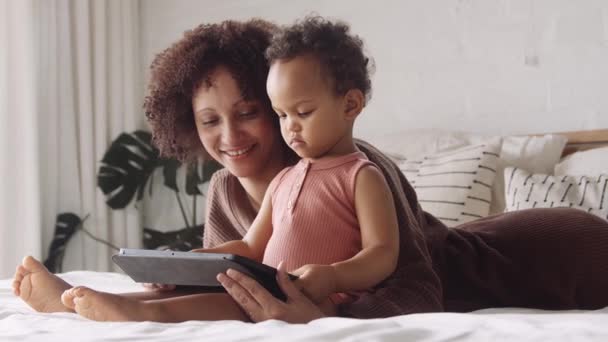 Madre Biracial y niño pequeño acostado en la cama en el dormitorio moderno, jugando en una tableta. Tecnología desde temprana edad. — Vídeos de Stock
