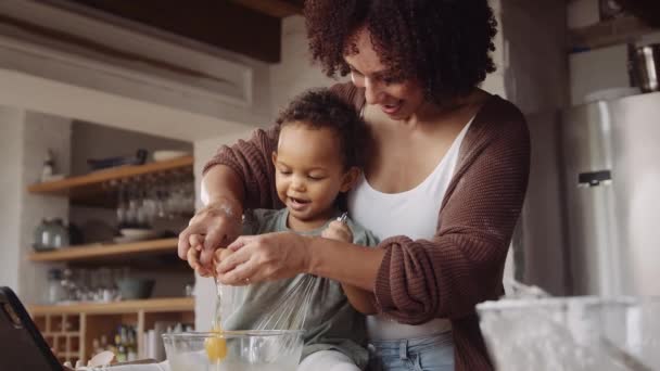 Biracial mother cracking egg in bowl in young daughters hands in modern-styled kitchen. Baking time with child. — Stock Video