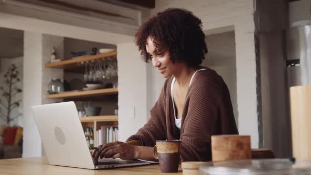 Innehåll och motiverad biracial mor som arbetar på laptop hemifrån i modernt kök. — Stockvideo