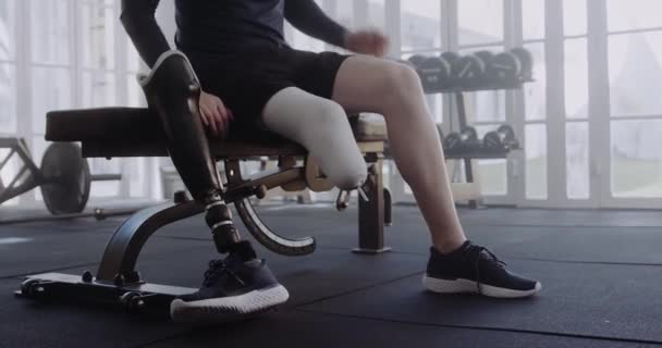 Biały mężczyzna siedzący na ławce i zakładający protezę nogi. Sportowiec niepełnosprawny ćwiczący w nowoczesnym stylu siłowni. — Wideo stockowe
