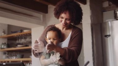 Çift ırklı anne ve küçük kız ellerinde unla el çırpıyorlar ve modern stil mutfakta pişiriyorlar.