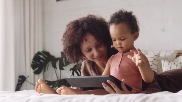Biracial anya bemutató kisgyermek tanulási tevékenységek tabletta, miközben feküdt az ágyban a hálószobában. — Stock videók