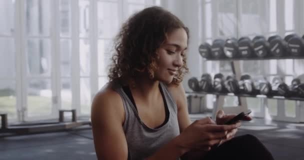 Biracial fena na podložce v moderní tělocvičně chystá cvičení. Usmívá se na svůj telefon, zatímco ona píše. — Stock video