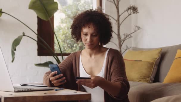 Biracial junge erwachsene Frau, die online mit dem Handy bezahlt, während sie tagsüber von zu Hause aus im modern gestylten Wohnzimmer arbeitet. — Stockvideo