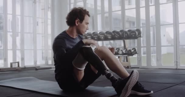 Běloch s protetickou koncovkou nohy sedí a zvedá mobil. Zdravotně postižený sportovec cvičí v moderní tělocvičně. — Stock video