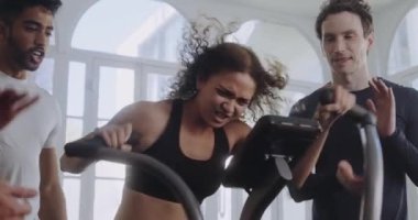 Modern stil bir spor salonunda çift ırklı bir kadın bisiklet üzerinde çok çalışıyor. Arkadaşları onu motive ediyor ve alkışlıyor..