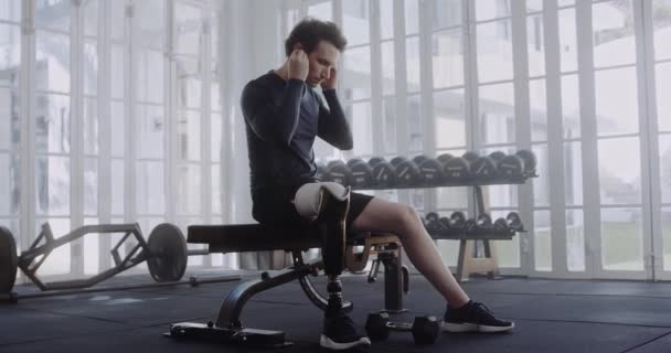 Белый мужчина с протезной ногой засовывает наушники в уши перед тренировкой. Спортсмен-инвалид, тренирующийся в современном спортивном зале. — стоковое видео