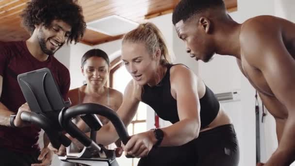 Zróżnicowana grupa młodych dorosłych, którzy wspierają białą przyjaciółkę podczas jazdy na stacjonarnym rowerze treningowym w siłowni. — Wideo stockowe