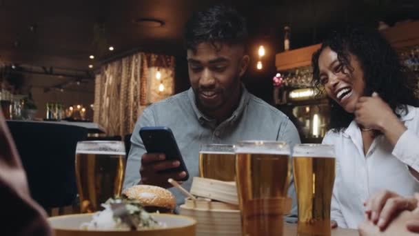 To multiraciale venner på en restaurant kigger på selfies de tog på en af deres mobiltelefoner. – Stock-video