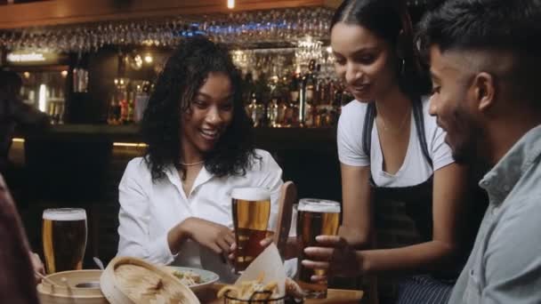 Çok ırklı garson, restorandaki bir grup arkadaşa biralarını getiriyor. Bir grup arkadaş bardaklarını tokuşturur.. — Stok video