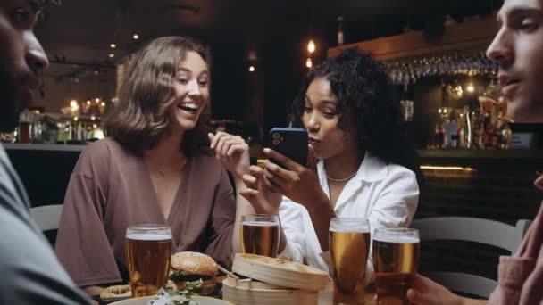 Dos felices mujeres multirraciales en un restaurante riéndose de algo en su teléfono móvil — Vídeos de Stock