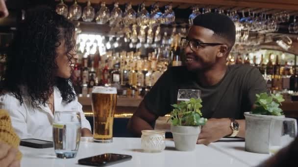 Bir restoranda arkadaşlarıyla birlikte çok ırklı bir adam ve kadın. Bira içiyorlar ve hoş bir sohbetin tadını çıkarıyorlar.. — Stok video