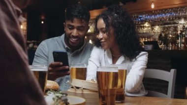 Bir restoranda cep telefonlarından birinin çektiği selfielere bakan iki çok ırklı arkadaş..