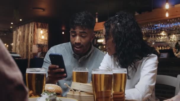 Dua teman multirasial di sebuah restoran melihat foto selfie yang mereka ambil dari salah satu ponsel mereka. — Stok Video