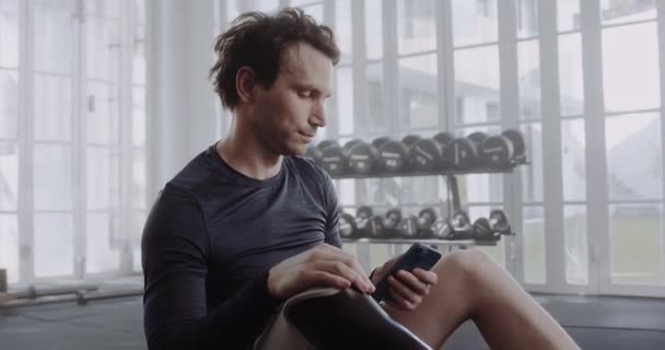 Caucasian male with prosthetic leg texting on his cellphone. Disabled athlete working out in modern-style gym. — Wideo stockowe