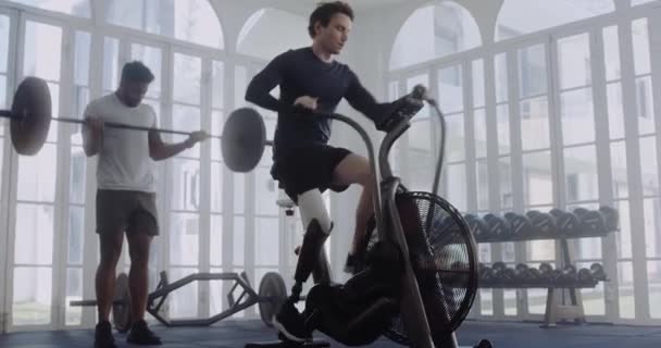 Καυκάσιος άνδρας με προσθετικό πόδι που ποδηλατεί με ποδήλατο αέρα ενώ ο σημαιοφόρος άνδρας σηκώνει βάρη στο βάθος. Αθλητής με ειδικές ανάγκες που γυμνάζεται σε σύγχρονο στυλ γυμναστήριο. — Αρχείο Βίντεο