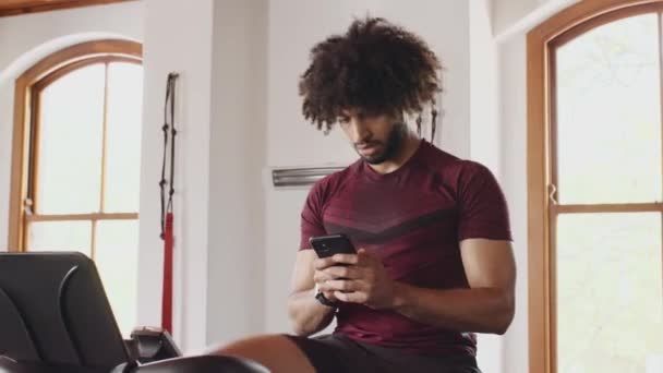 Young adult Middle Eastern male cycling on indoor fitness bike. Concentrating on texting on cellphone in an indoor fitness gym. — Stock video