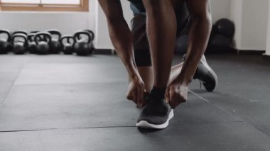 Slow motion video of fit and healthy black male confidently kneeling down and tying his shoe lace at an indoor fitness gym