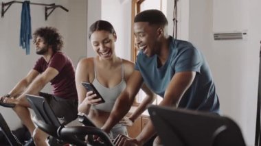 Çift ırklı kadın spor salonundaki spor salonunda siyah bir erkeğin komik görüntüsünü cep telefonuyla gösteriyor.