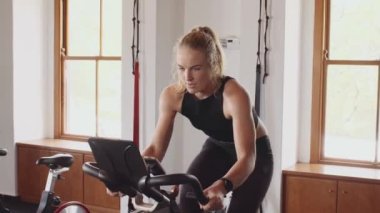 Kapalı spor salonunda spor bisikleti süren kararlı, genç, beyaz bir kadın. Kendine güven ve sağlık.