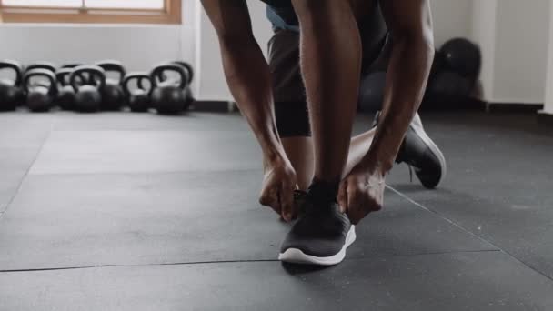 Slow motion video of fit and healthy black male confidently kneeling down and tying his shoe lace at an indoor fitness gym — Vídeo de Stock