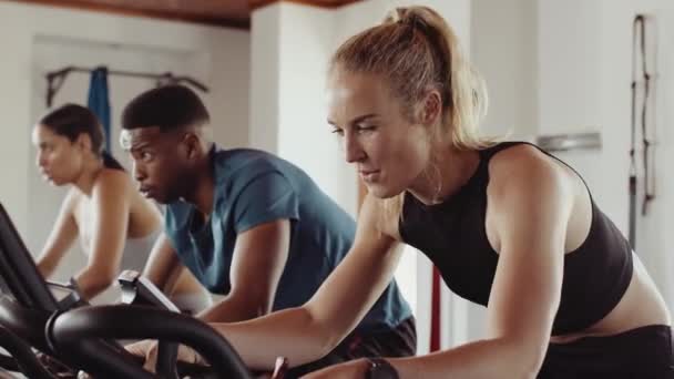 Caucasian female cycling on stationary bike in a group fitness class at an indoor fitness gym. Having a good time and determined. — Stok video