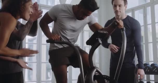 Biracial male reaching his personal best and working hard on the bike in a modern style gym. Friends motivating, cheering him on. — Stock video