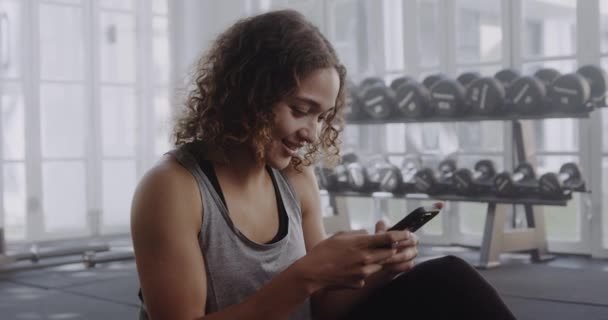 Biracial female sitting on mat at modern gym about to do workout. Smiling at her phone while she texts. — стоковое видео