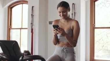 Cep telefonuyla mesajlaşırken gülümseyen genç, çift ırklı bir kadın. Spor salonunda sabit egzersiz bisikletinde oturmak..