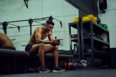 Genç Afro-Amerikalı bir erkek çevrimiçi bir mesaja cevap vermek için telefonuna bakıyor. Spor salonunda otururken telefonuna gülümseyen melez bir erkek. Yüksek kalite fotoğraf