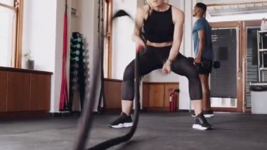 Kararlı genç beyaz kadın, kapalı spor salonunda güç kazanmak için savaş ipleri kullanıyor..