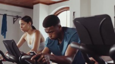 Odaklanmış siyah erkek ve çift ırklı kadın sabit bisiklete biniyor. Spor salonunda eğleniyor ve kararlı.. 