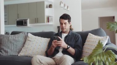 Caucasian man relaxing sitting on couch at home while typing a message on his mobile phone. High quality 4k footage