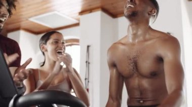 Diverse group of fit young adults supporting their black male friend while he cycles on stationary exercise bike at indoor fitness gym. 