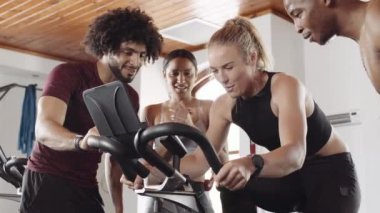 Diverse group of fit young adults cheering around Caucasian female friend while she cycles on stationary exercise bike at indoor fitness gym. 