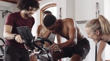 Diverse group of fit young adults cheering around black male friend while he cycles on stationary exercise bike at indoor fitness gym. 