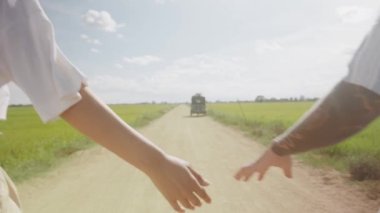 Close-up of young couple holding hands while walking away on dirt road through agricultural fields