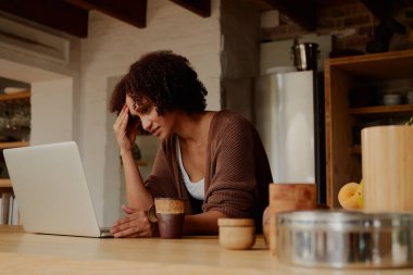 Frustrated young biracial woman in casual clothing working on laptop in kitchen at home